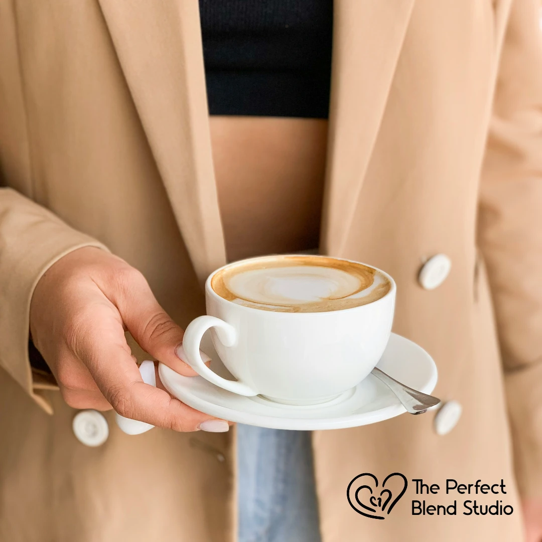 A person in a beige blazer holds a white cup of latte with a swirl design, on a matching saucer with a tiny spoon.