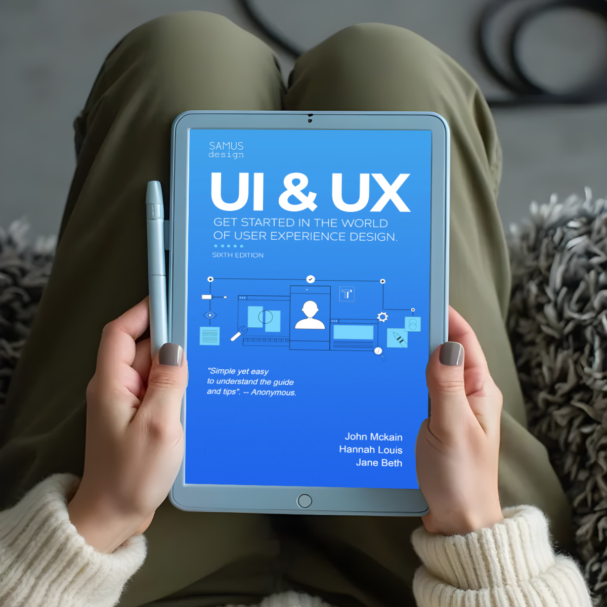 Tablet displaying UI and UX design book cover. Blue background with white text. Person holding device, wearing cream sweater and olive pants. Stylus visible. Cozy setting on textured rug.
